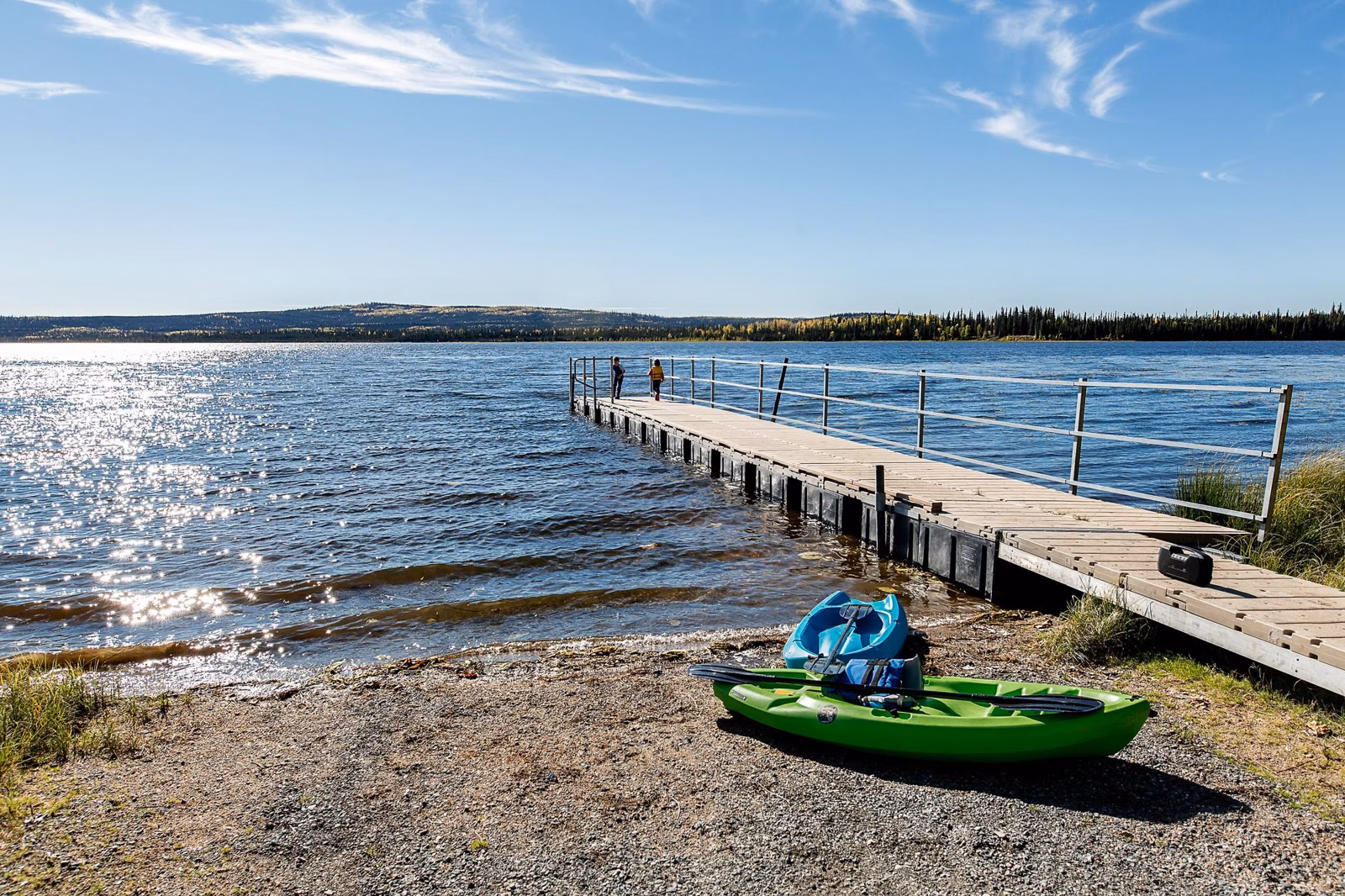 A green kayak pulled up on a gravel lake shore beside a wooden dock on a sunny day