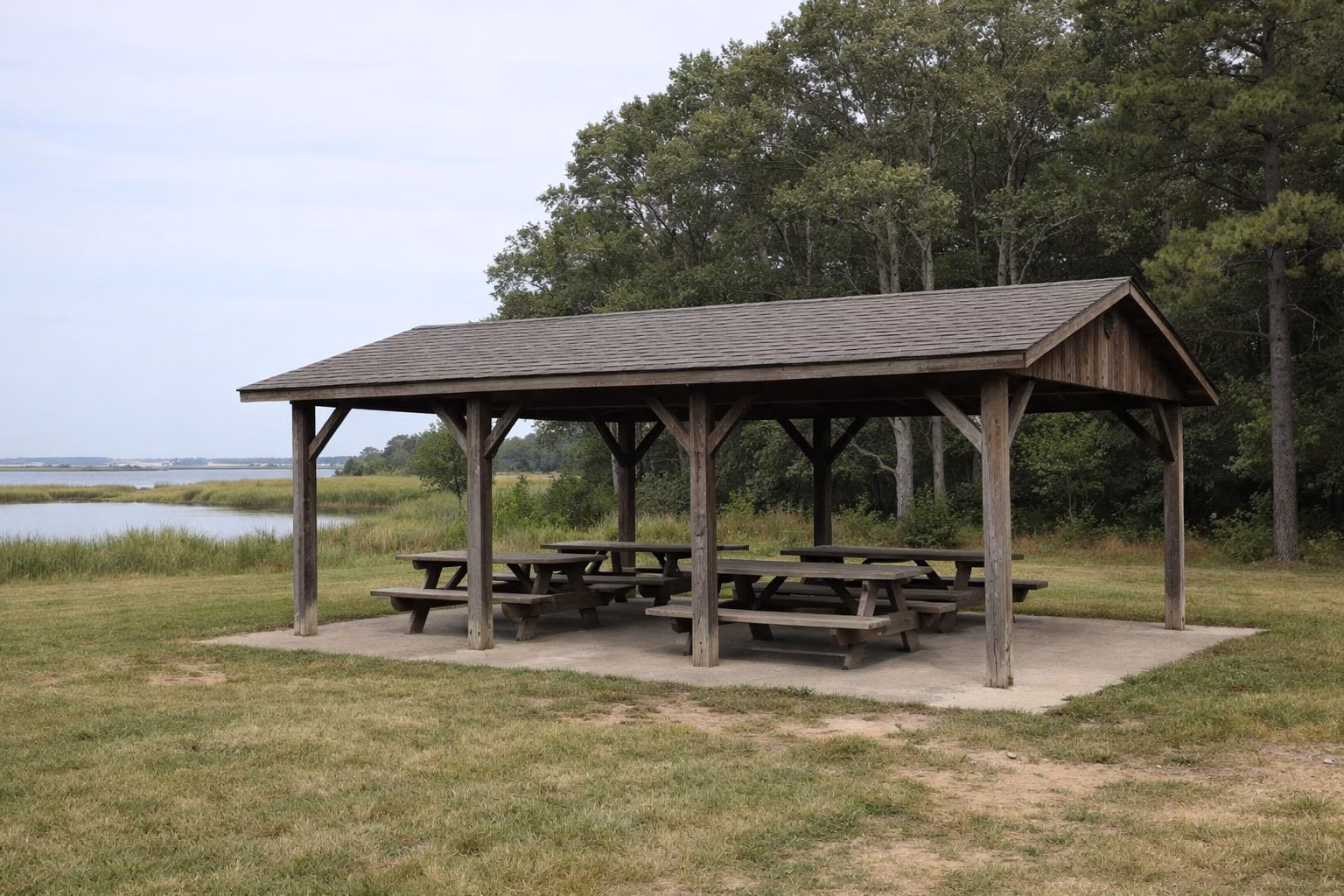 An open-sided wooden picnic shelter with picnic tables at the edge of a mown grass field, oak woods behind