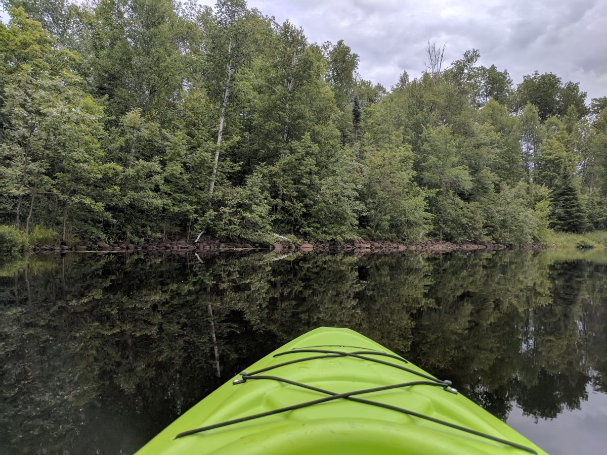 The bow of a green kayak on still water reflecting a wooded shoreline